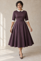 Aubergine back-button raglan dress design preview on an adult woman, front view showing the ivory rounded collar, diagonal raglan joins and smooth flared pocket skirt.
