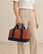 Design preview: adult woman holding both navy handles of the same compact rust duffel beside her hip