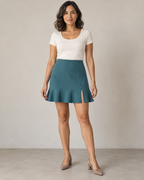 Front design preview of a muted petrol flounce-hem mini skirt on an adult woman, with a fitted faced waist and short left-front slit